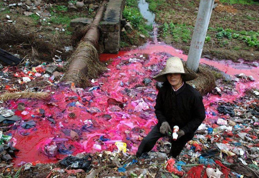 Žena zbiera plastové fľaše z rieky, ktorá je znečistená fabrikou a má ružovú farbu.